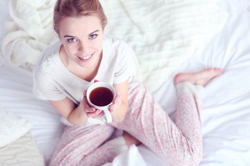 Attractive young blonde girl in a pajamas holding a cup of tea while sitting in white bedding and pillows