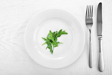 Arugula leaves in plate with fork and knife on wooden table, top view