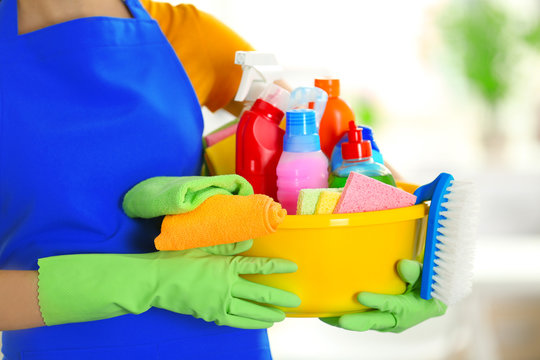Woman in rubber gloves holding basin with detergents on blurred background