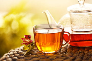 Glass cup of tea with teapot on wicker mat against blurred background