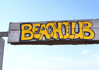 Beach club signboard with yellow letters on a wooden plank. Entrance sign of a beach restaurant or Beachclub. Close-up with blue sky. 