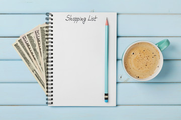 Coffee mug and notebook with shopping list and cash money dollar on blue rustic table from above