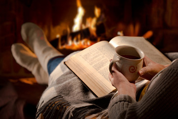 Woman resting with cup of hot drink and book near fireplace