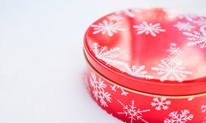 Christmas gift container.  Red, round cookie & baked goods tin container decorated in red with white snowflake print pattern, sitting in natural snow.