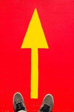 Two feet in sneakers standing behind yellow traffic arrow road sign on the red street asphalt background 