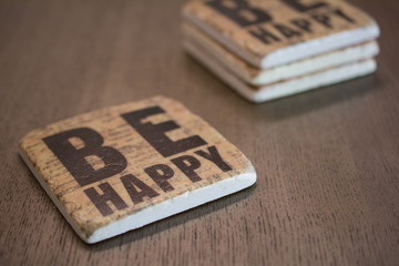 A set of stone coasters stacked on a wooden table surface reading Be Happy