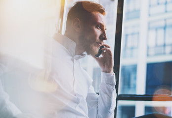 Photo bearded adult businessman working on modern loft office. Man wearing white shirt and using contemporary smartphone. Panoramic windows background. Horizontal, film effect, bokeh