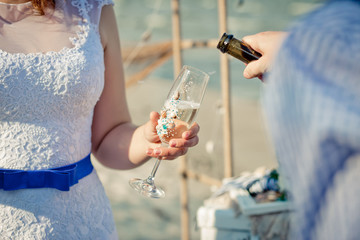 Glass with champagne is  in the bride's hands. The groom is pouring champagne into the bride's glass.  Sea style wedding.