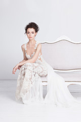 Beautiful young bride in a luxurious lace wedding dress. She sits on a white vintage sofa. She is an elegant and slim. The dress has a translusent silk train. Her hands are folded in elegant gesture.