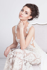 Portrait of beautiful young bride in a luxurious lace wedding dress. She sits on a white vintage sofa. She is an elegant and slim, she has tannes skin, dark hair and gray eyes