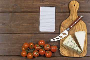 Cheese with white mold it and stainless steel cheese knife, cherry tomatoes and paper notebook are lying on a wooden board.