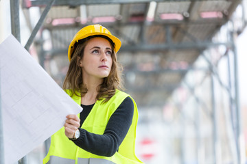 Female construction worker with helmet and safety jacket on construction site examining office...