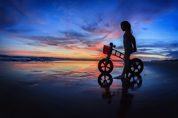 Fototapeta premium Black silhouette of little child - run bike rider stand on wet sand beach, look at sea surf and colourful sunset sky. Family travel lifestyle background, outdoor fun activity on Bali summer vacation.