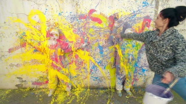 Time lapse of two street artists creating a colorful abstract mural by splashing acrylic paints on a wall and two models in protective coveralls to leave white silhouettes on multi-colored wall