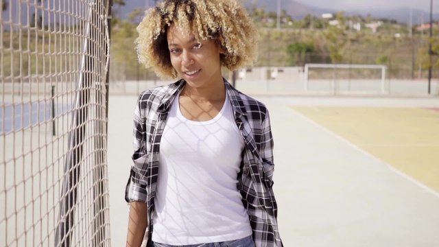 One pretty young black woman with skateboard in hand wearing jean shorts and unbuttoned plaid top poses near ball court under a bright sun