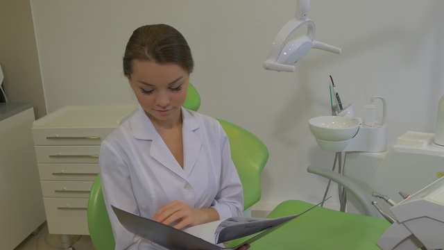 Dantist is Reading a Notes in a Fold Raises Head Girl in Lab Coat is Smiling Sitting on a Chair at Dental Treatment Room Young Pretty Doctor Woman