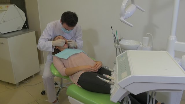 Young Man Dentist in Mask is Treating a Teeth Examining a Teeth of a Patient at Dental Treatment Room Young Woman is Lying on a Green Chair Napkin on Chest