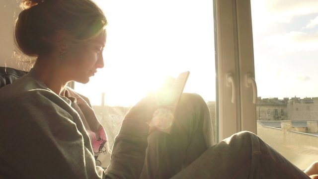 beautiful young woman sitting on window sill with coffee end reading book on background of sunset