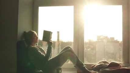 beautiful young woman sitting on window sill with coffee end reading book on background of sunset - Powered by Adobe