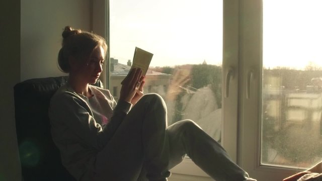 beautiful young woman sitting on window sill with coffee end reading book on background of sunset