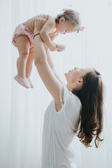 Beautiful mother portrait playing with her cute little girl in a white room in the morning