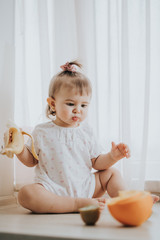 Cute little baby girl on the table in the kitchen eats citrus fruits full of vitamins and makes funny cute faces