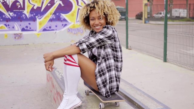 Trendy young woman sitting on a skateboard waiting for someone in an urban environment with graffiti