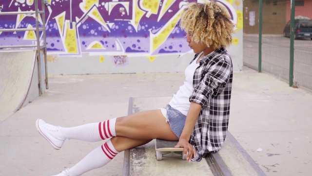Trendy young woman sitting on a skateboard waiting for someone in an urban environment with graffiti