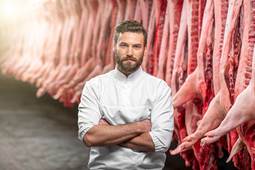 Portrait of a handsome butcher in white uniform at the meat manufacturing with pork carcasses on the background