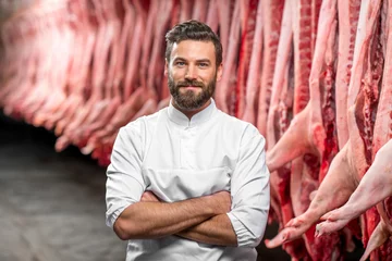 Fotobehang Portrait of a handsome butcher in white uniform at the meat manufacturing with pork carcasses on the background © rh2010