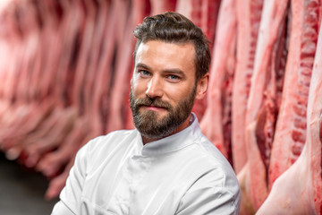 Portrait of a handsome butcher in white uniform at the meat manufacturing with pork carcasses on...