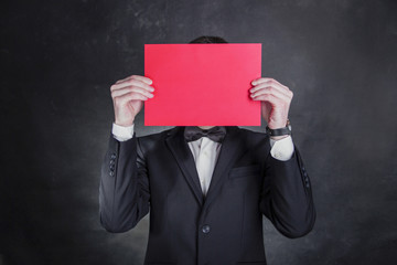 Man wears black tuxedo holds paper in front of face.