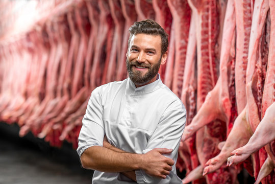 Portrait of a handsome butcher in white uniform at the meat manufacturing with pork carcasses on the background