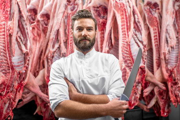 Portrait of a handsome butcher holding knife standing on the pork carcasses background at the meat manufacturing