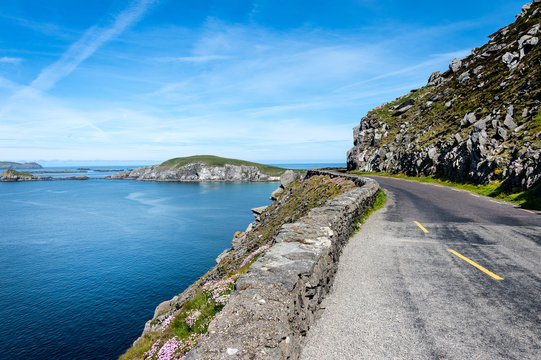 Ireland, Dingle, Slea Head: Street scene with panoramic view over coast, ocean water, bending empty street, the hill of Slea Head and blue sky in the background