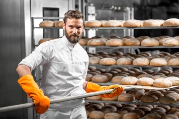 Tuinposter Handsome baker in uniform with orange working gloves putting with shovel from the oven bread loafs on the shelves at the manufacturing © rh2010