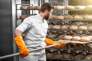 Handsome baker in uniform with orange working gloves putting with shovel from the oven bread loafs on the shelves at the manufacturing - obrazy, fototapety, plakaty