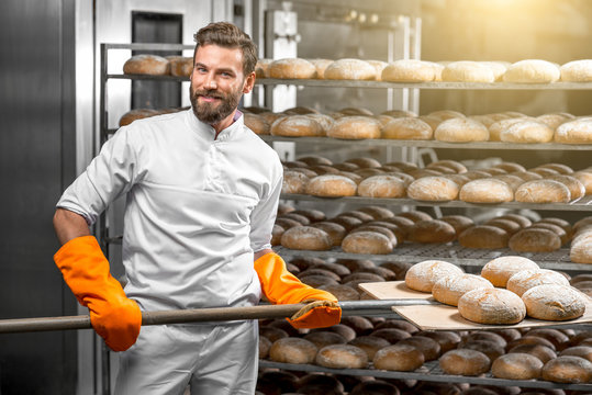 Handsome baker in uniform with orange working gloves putting with shovel from the oven bread loafs on the shelves at the manufacturing