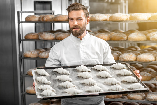 Handsome baker in uniform holding tray full of freshly baked croissants at the manufacturing