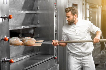 Handsome baker in uniform taking out with shovel freshly baked buckweat bread from the oven at the manufacturing - obrazy, fototapety, plakaty