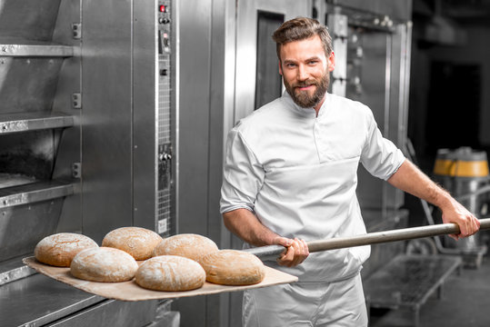 Handsome baker in uniform taking out with shovel freshly baked buckweat bread from the oven at the manufacturing