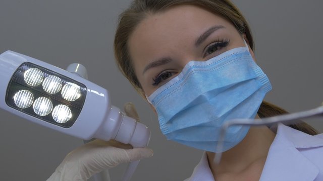 Woman Dentist in Mask is Holding a Dental Hook Standing Upon a Patient Looking at Camera Doctor in Lab Coat Hands in Gloves Examining a Teeth Dental Clinic