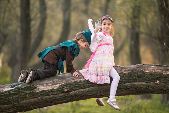 Portrait of a cute little girl dressed up as a fairy and waving while sitting on a tree in a forest with a boy in knight costume
