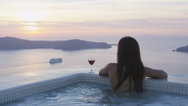Luxury travel lifestyle woman in pool at sunset. Young woman having glass of red wine in swimming pool. Asian female is watching sunset over sea. She is enjoying her vacation on Santorini, Greece.