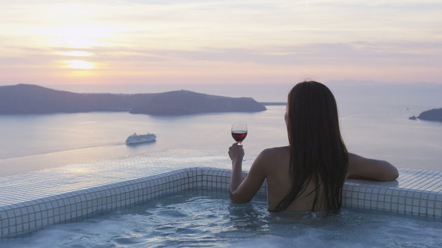 Jet set Travel woman in bikini in pool by amazing sunset view. Model placing glass of red wine at the edge of swimming pool. Asian female is enjoying her vacation on Santorini, Greece. RED EPIC.