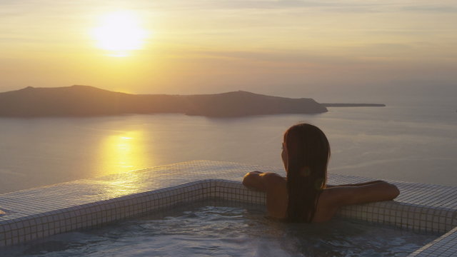 Young woman relaxing at the edge of swimming pool. Female is watching beautiful view of sunset. She is enjoying her vacation at resort by sea. Santorini, Greece.