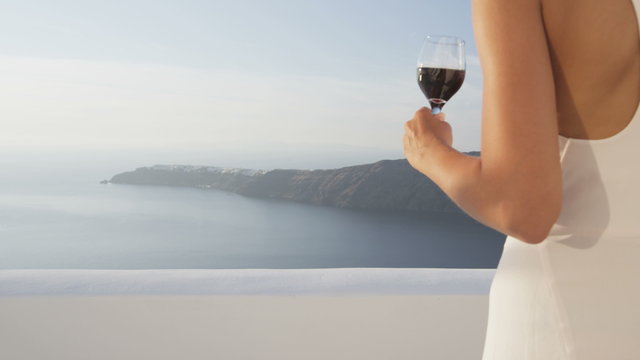 Elegant young woman holding red wine glass outside walking at terrace in luxury villa enjoying beautiful sea view and mountains. Female in white sundress on summer vacation travel. Santorini, Greece.