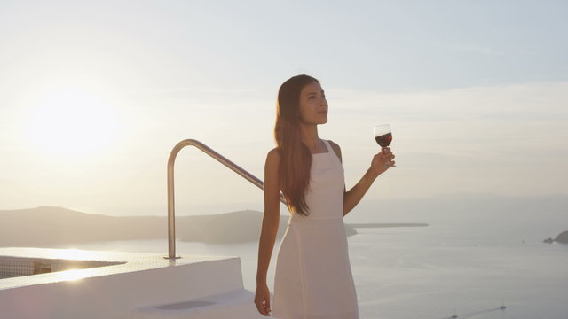 Attractive woman with glass of red wine walking down steps of swimming pool. Female is at resort by sea during sunset. She is in her white sundress during vacation.