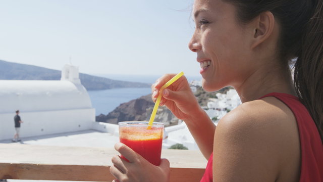 Happy young woman drinking fresh red beet and carrot juice. Healthy female enjoying drink at outside cafe looking at church and landscape during summer vacation on Santorini, Greece.