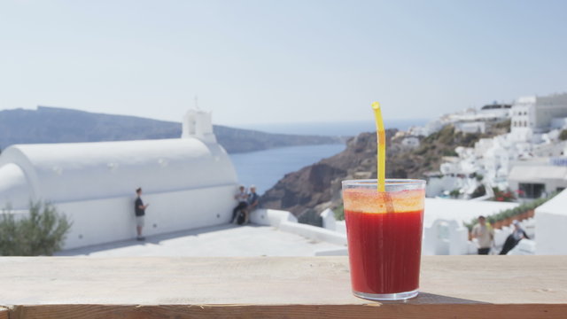 Healthy juice glass on railing at observation point. Organic beet and carrot juice. High angle view of tourists visiting church during summer. View of sea landscape anad clear sky on Santorini, Greece